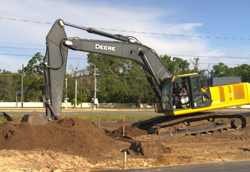John Deere tracked excavator digging a utility trench beside a stockpile of excavated soil at a Tampa Bay job site, with survey stakes marking the line and a roadway behind it