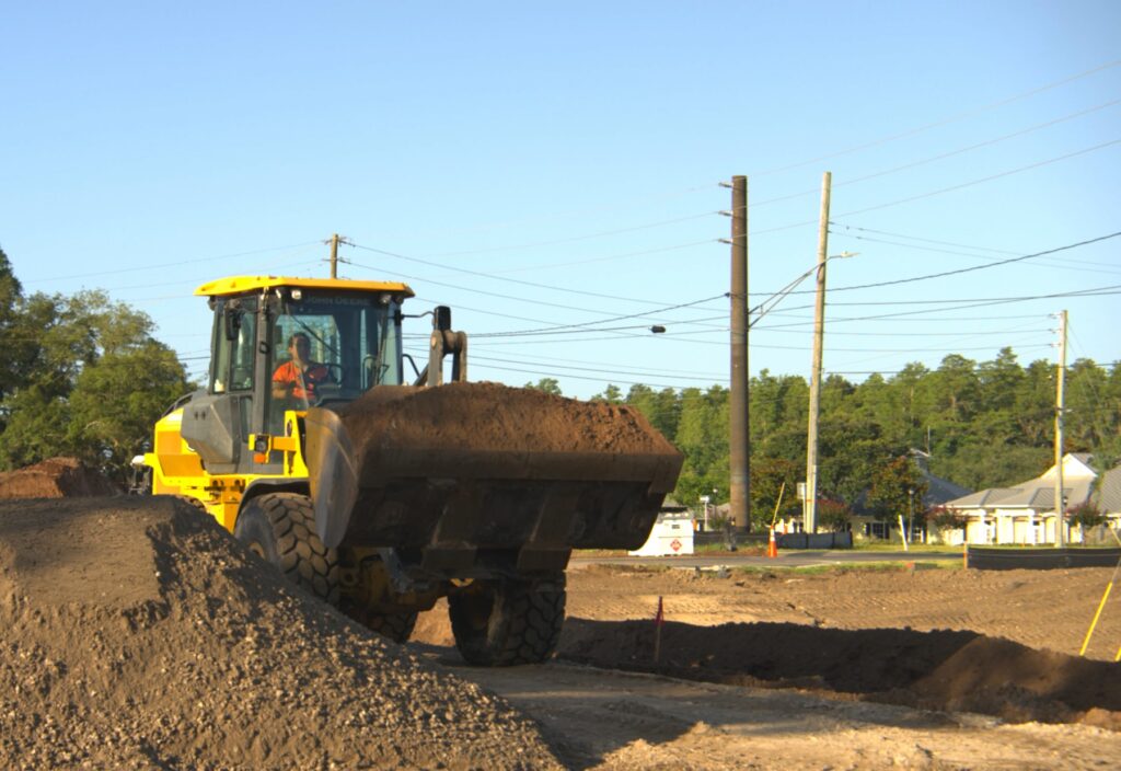 John Deere wheel loader carrying a full bucket of excavated dirt up a soil stockpile at a Tampa Bay site preparation job, with a graded pad, utility poles and a fuel tank behind it